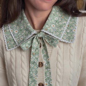May include: A light green floral collar with a white lace trim and a matching bow. The collar is paired with a cream-colored cable knit cardigan with brown buttons. The floral pattern features small white daisies.