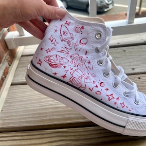 Può includere: Sneaker alta bianca con illustrazioni rosse a tema spaziale, tra cui un razzo, pianeti e stelle. La scarpa ha una striscia nera lungo la suola e lacci bianchi. La sneaker è su una superficie di legno.