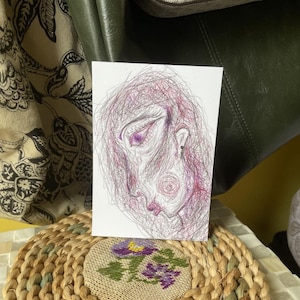 May include: A small, white card with a pen and ink drawing of a face in profile. The artwork is in purple and pink, with a scribbled texture. The face has a prominent eye and a small earring. The style is reminiscent of a sketch.