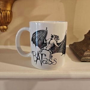 Peut inclure: Mug en céramique blanche avec une illustration en noir et blanc d'une chauve-souris aux ailes déployées. Le mot "BATass" est imprimé en dessous. La tasse a une anse incurvée.