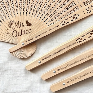 May include: Wooden hand fans with intricate cut-out designs. One fan displays the text "Mis Quince" with a butterfly and crown. The handles are engraved with "Riley's Quinceañera 06.15.2028".