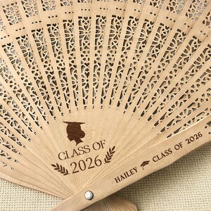 May include: A light-colored wooden fan with intricate cut-out designs. The fan is engraved with "CLASS OF 2026" and a graduation cap silhouette. The name "Hailey" is also engraved, along with "CLASS OF 2026".