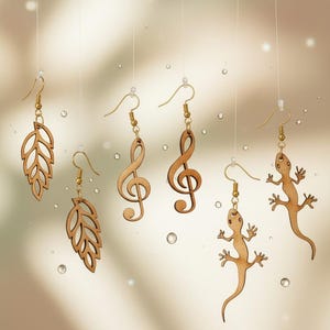 Puede incluir: Una colección de pendientes de madera con ganchos dorados. Los pendientes presentan varios diseños, incluyendo formas de hojas, claves de sol y figuras de lagartos. Los pendientes están suspendidos sobre un fondo suave y borroso.