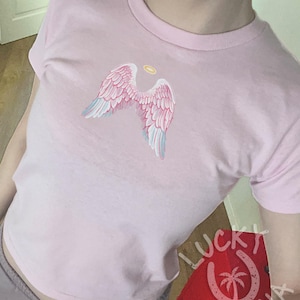 Op de afbeelding: Lichtroze T-shirt met een ontwerp van roze en lichtblauwe engelen vleugels en een gouden halo. De halslijn is rond. Het Lucky Laguna-logo is rechtsonder zichtbaar. Casual stijl.