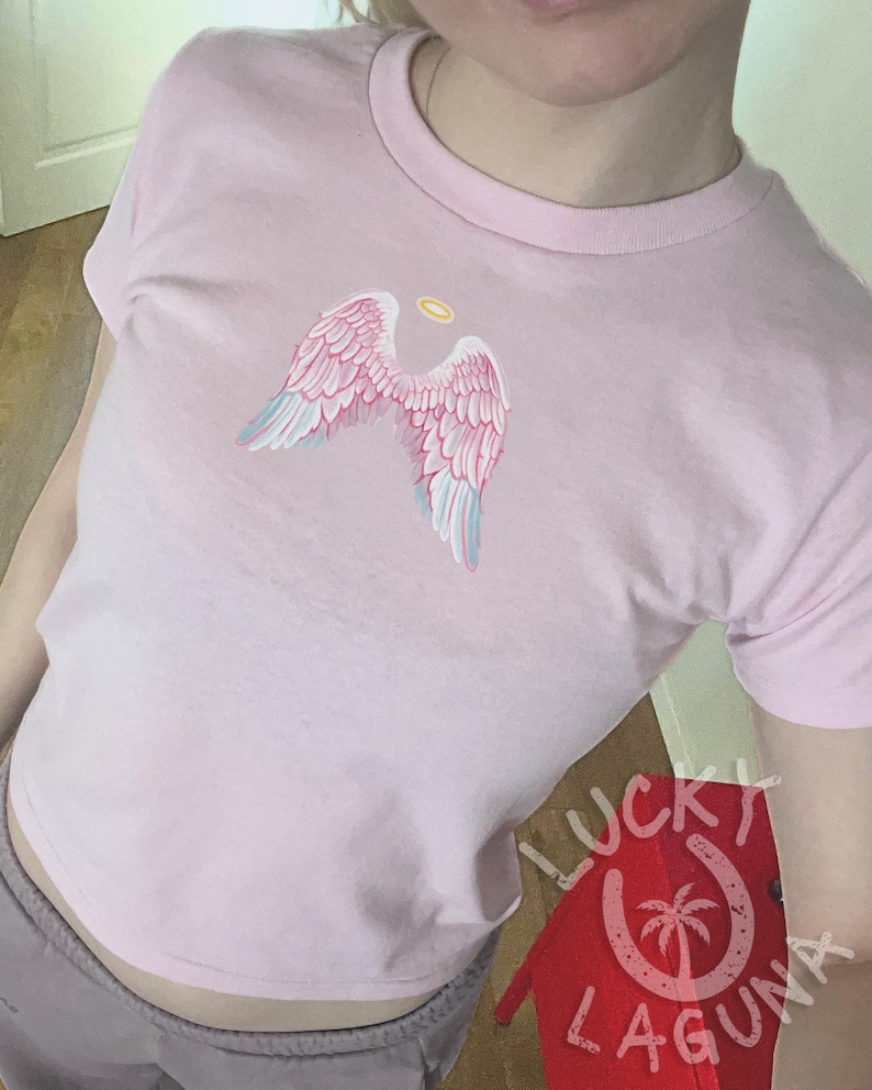 Op de afbeelding: Lichtroze T-shirt met een ontwerp van roze en lichtblauwe engelen vleugels en een gouden halo. De halslijn is rond. Het Lucky Laguna-logo is rechtsonder zichtbaar. Casual stijl.