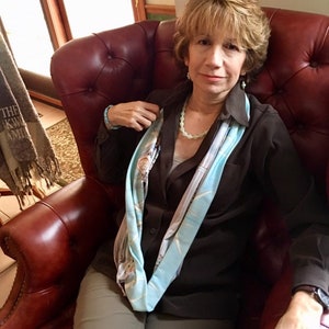 May include: A woman wearing a brown shirt with a blue and white scarf, sitting in a red leather armchair. She is wearing a necklace and bracelets.