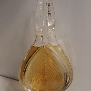 Könnte beinhalten: Eine transparente Parfümflasche aus Glas mit einem einzigartigen, blattartigen Design. Die Flasche ist mit einer goldenen Flüssigkeit gefüllt und hat einen transparenten Verschluss mit dem Wort "CHAMADE" in Gold. Auf der Basis der Flasche steht das Wort "GUERLAIN".