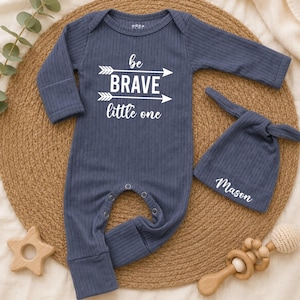May include: A dark blue ribbed long-sleeve romper with a white graphic print that reads "be BRAVE little one" between two arrows. A matching dark blue ribbed beanie with the name "Mason" printed in white is positioned next to the romper.