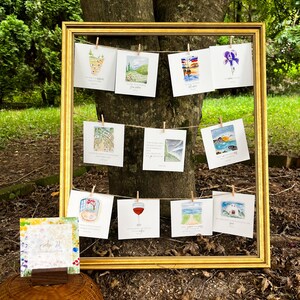 May include: A gold-framed display featuring watercolor art prints clipped to a string with small wooden clothespins. The prints depict various landscapes, a wine glass, and a house. A small card with floral designs and text is in the foreground.
