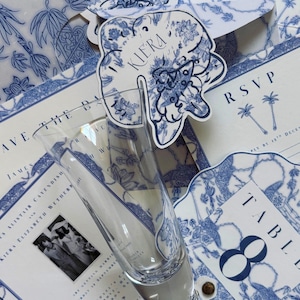 Könnte beinhalten: Eine Auswahl an Hochzeits-Papeterie mit blauem und weißem Blumendesign. Enthält eine Sektflöte, Einladungskarten, RSVP-Karten und eine Tischkarten. Der Name "Kiera" ist auf einem dekorativen Anhänger aufgedruckt.