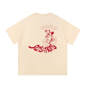 May include: A cream-coloured short-sleeve t-shirt featuring a red graphic print on the back. The design depicts a person riding a bicycle with the word GOOD arched above and the word SENSATIONS written in a wavy, bold red font below the bicycle.