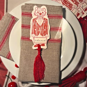 Puede incluir: Un cubierto con una servilleta de lino gris con rayas rojas, un plato blanco y una tarjeta de nombre decorativa. La tarjeta de nombre es una ilustración de dibujos animados de un gato con traje de rayas, con una borla roja. El nombre "Edward" está escrito en la tarjeta.