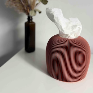 May include: A round, terracotta-coloured tissue box cover with a vertical ribbed texture. The container has a wide circular opening at the top, from which a single white paper tissue is protruding. The object has a smooth, matte finish.