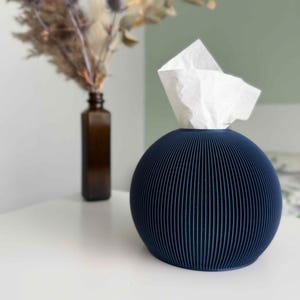 May include: A spherical, dark blue tissue box cover featuring a vertically ribbed, pleated texture. A single white tissue is pulled through the top opening of the round container, which sits on a flat surface next to a brown glass bottle.