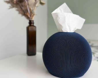 May include: A spherical, dark blue tissue box cover featuring a vertically ribbed, pleated texture. A single white tissue is pulled through the top opening of the round container, which sits on a flat surface next to a brown glass bottle.