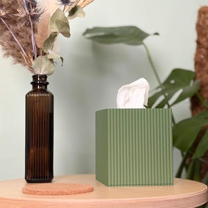 May include: A square, sage green tissue box cover with a vertical ribbed texture. A single white tissue protrudes from the top opening. The box is positioned next to a tall, amber-coloured glass bottle containing dried flowers and a round cork coaster.