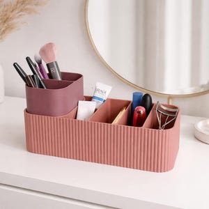 May include: A two-tiered dusty rose makeup organiser with a ribbed exterior texture. The unit features a raised back section for brushes and a lower front section with multiple vertical dividers for storing various cosmetics and beauty tools.
