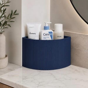May include: A dark blue, semi-circular corner storage organiser with a vertical ribbed texture. Inside the organiser are three skincare bottles: a white tube labelled The Ordinary, a white pump bottle labelled CeraVe, and a beige bottle labelled OUAI.