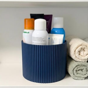 May include: A dark blue cylindrical storage container with a vertical ribbed texture, holding various skincare bottles and tubes with white, orange, and blue caps. The container is positioned on a white shelf next to two rolled beige towels.