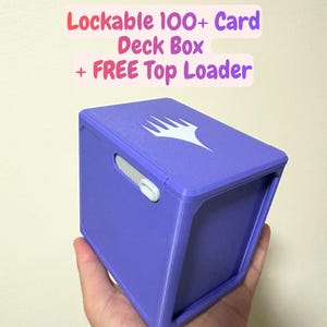 May include: A purple rectangular deck box with a white logo on top and a white sliding lock mechanism on the side. The text above the box reads: Lockable 100+ Card Deck Box + FREE Top Loader. The box is designed to hold over 100 playing cards.