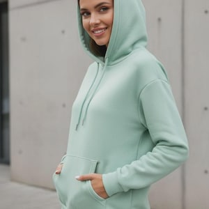 May include: A light green hoodie with a drawstring hood and a front kangaroo pocket. The hoodie is a solid color and appears to be made of a soft material. The model is wearing blue jeans.