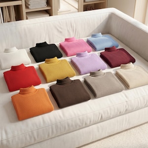 May include: A collection of folded turtleneck sweaters in various colors, including white, black, pink, and blue, arranged on a white sofa. Other colors include red, yellow, purple, burgundy, tan, orange, and brown. The sweaters are made of a ribbed knit material.
