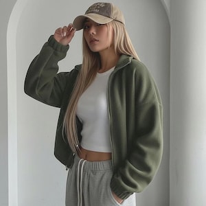 May include: An olive green fleece zip-up jacket with a white crop top and gray sweatpants. A tan and brown baseball cap is worn. The outfit is casual and comfortable.