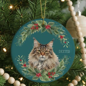 May include: A round, teal Christmas ornament featuring a detailed portrait of a cat with brown and tan fur. The cat is surrounded by a wreath of green pine needles and red berries. The name "DEXTER" is printed on the right side of the ornament.