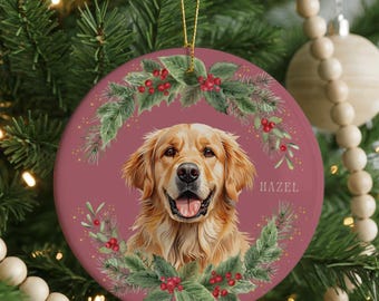 May include: A round Christmas ornament featuring a Golden Retriever dog with a happy expression, surrounded by a wreath of green leaves and red berries. The dog's name, "HAZEL", is printed on the right side. The ornament hangs from a gold string.