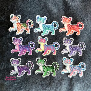 May include: Nine individual stickers of cartoon tiger cubs arranged in a grid. Each cub features a unique colour palette, including rainbow, teal, orange, pink, multicoloured, purple, violet, green, and pastel pink and blue stripes with white accents.