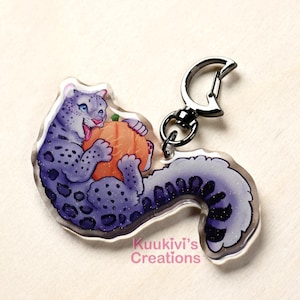 Peut inclure: Un porte-clés en acrylique transparent représentant un léopard des neiges violet et gris tenant une citrouille orange. Le porte-clés a un fermoir argenté et le texte "Kuukivi's Creations". Le léopard des neiges a les yeux bleus et sourit.