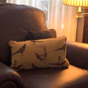 Könnte beinhalten: Ein rechteckiges Zierkissen mit hellgelbem Hintergrund, verziert mit Illustrationen von Fasane in Braun, Beige und Schwarz. Das Kissen liegt auf einem braunen Ledersessel, mit einem kleineren karierten Kissen dahinter.