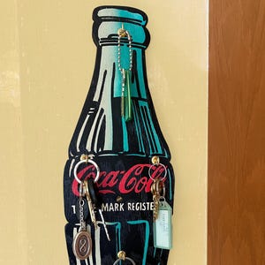 Könnte beinhalten: Ein hölzerner Schlüsselhalter in Form einer Coca-Cola-Flasche. Der Schlüsselhalter ist schwarz mit weißen und türkisfarbenen Akzenten und zeigt das Coca-Cola-Logo in Rot. Mehrere Schlüssel und Schlüsselanhänger hängen daran.