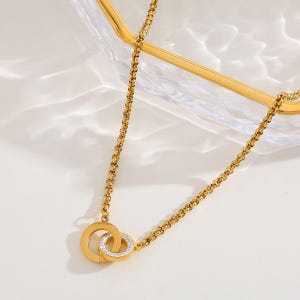 May include: A gold-toned chain necklace featuring two interlocking circular pendants. One pendant is a solid gold-toned ring, while the other is a gold-toned ring encrusted with small, clear, sparkling stones along its outer surface.