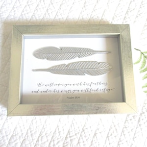May include: A framed silver metal wall art with two feathers and a bible verse from Psalm 91:4, "He will cover you with his feathers, and under his wings you will find refuge."