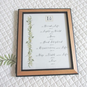 May include: A framed print with a white background and a green leafy vine on the left side. The text reads "I AM" in a square box at the top, followed by a list of phrases from the Bible, including "Bread of Life", "Light of the World", "Door", "Good Shepherd", "Resurrection and the Life", "Way, Truth and the Life", and "the Vine".