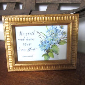 May include: A gold-colored framed print featuring a watercolor floral arrangement of blue hydrangeas. The print includes the text "Be still and know that I am God." from Psalm 46:10.