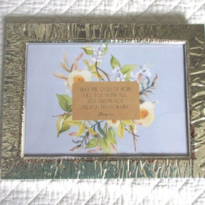 May include: A framed print with a floral design and a gold-colored textured frame. The print features a watercolor-style bouquet with white and yellow flowers, green leaves, and the text "MAY THE GOD OF HOPE FILL YOU WITH ALL JOY AND PEACE AS YOU TRUST IN HIM."