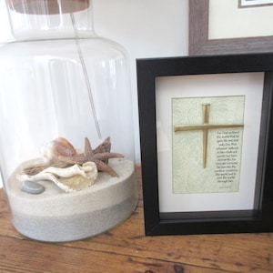 May include: A framed print featuring a gold nail cross and a Bible verse. The frame is black and the background of the print is green.