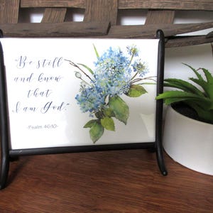May include: A white ceramic tile with a black metal stand. The tile features the text "Be still and know that I am God" and a watercolor painting of blue hydrangeas. A potted plant is in the background.