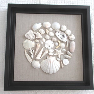 May include: A framed artwork featuring a collection of various white seashells, arranged in a circular pattern on a beige linen background. The black frame surrounds the seashells, creating a coastal theme. Some shells have a pink hue, and one shell has the text "Be still and know..."