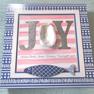 May include: A framed wall decor with a pink and white striped background. The word "JOY" is spelled out in metal letters with a seashell in the centre. The bottom of the frame has the text "Jesus first, then Others, Yourself last".