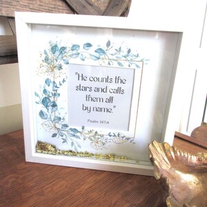 May include: White framed art print with the quote "He counts the stars and calls them all by name." Floral and leaf accents in blue and gold surround the text. Psalm 147:4 is printed below the quote.