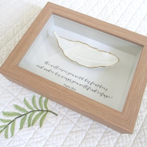 May include: A framed decorative piece featuring a white feather-shaped dish with gold trim. The frame is light brown, and the background includes text from Psalm 91:4. A green embroidered fern is in the lower left corner.