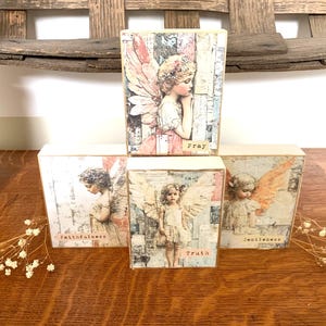 May include: Four decorative wooden blocks with angel illustrations. Each block features a different image of an angel with text: "Pray", "Faithfulness", "Truth", and "Gentleness". The blocks are on a wooden surface.