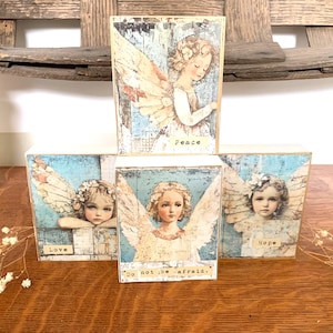 May include: Four wooden blocks with angel illustrations. Each block features a different image of an angel with wings, and the words "Peace", "Love", "Hope", and "Do not be afraid" are printed on the blocks. The blocks are arranged on a wooden surface.