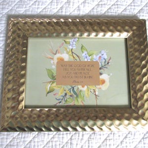 May include: A rectangular gold-colored picture frame with a textured surface. Inside the frame is a floral print with a quote on a tan background. The quote reads: "MAY THE GOD OF HOPE FILL YOU WITH ALL JOY AND PEACE AS YOU TRUST IN HIM."