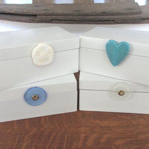 May include: Four white wooden boxes with different decorative knobs. The boxes are stacked on top of each other. The knobs are a seashell, a heart, a blue circle, and a clear circle.