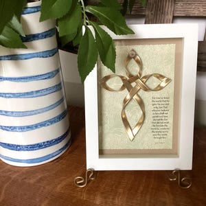 May include: A white frame containing a gold Celtic knot cross. The frame is on a wooden surface and features a quote about God's love for the world. "For God so loved the world that he gave his one and only Son, that whoever believes in him shall not perish but have eternal life. For God did not send his Son into the world to condemn the world, but that the world through him might be saved." - John 3:16-17