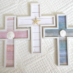 May include: Three wooden crosses with white frames. The cross on the left has a pink and white woodgrain design with a white seashell in the center. The cross in the middle has a white woodgrain design with a gold starfish in the center. The cross on the right has a blue and white woodgrain design with a white seashell in the center.